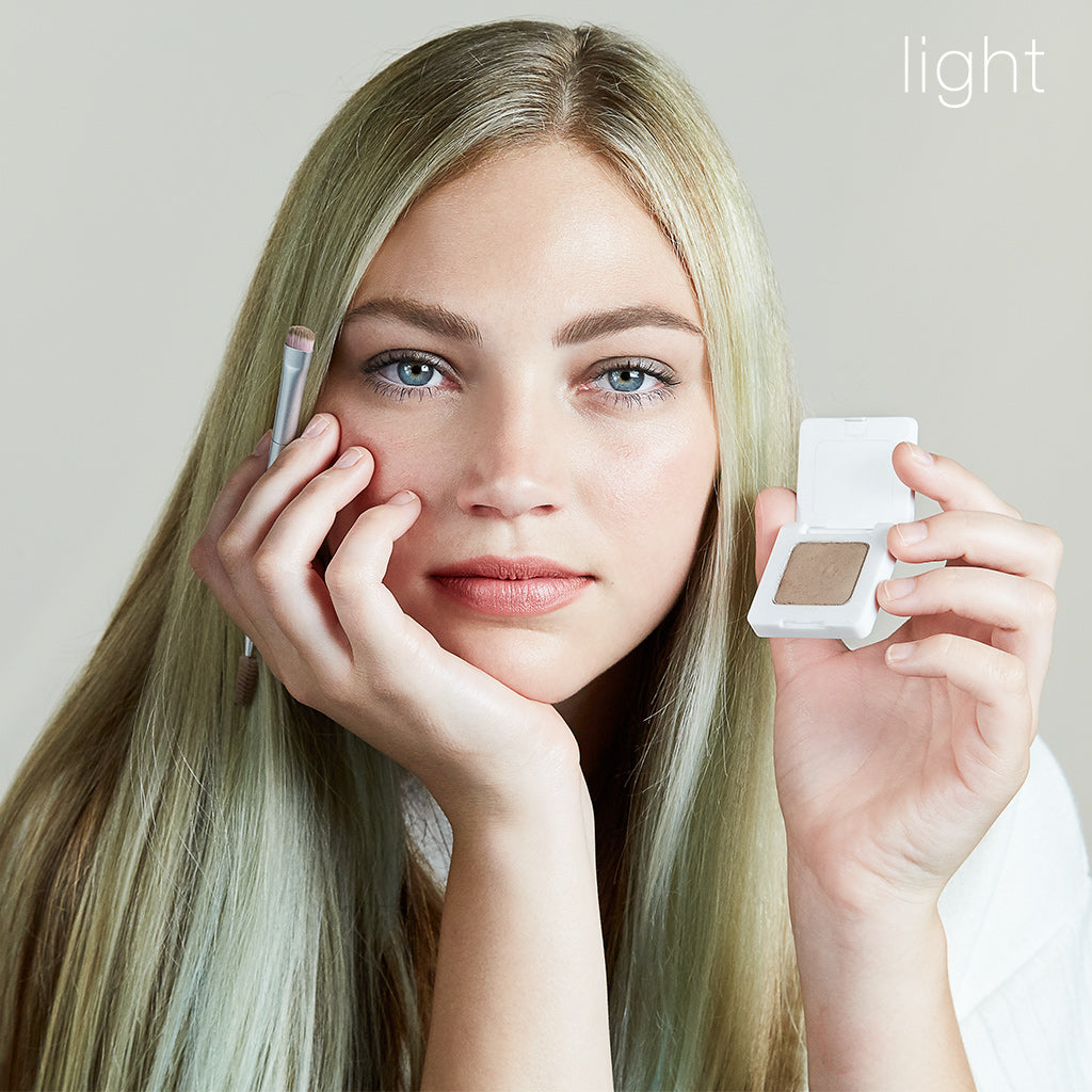 Model wearing Back2Brow Powder in shade Light, holding Back2Brow Brush in right hand, holding Back2Brow Powder outer carton product in left hand